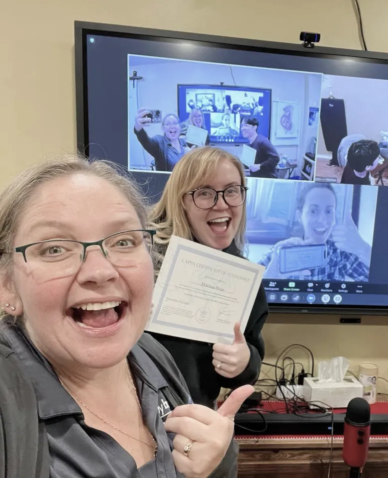 Two women posing with a certificate in front of a Vibe Board showing a video meeting.