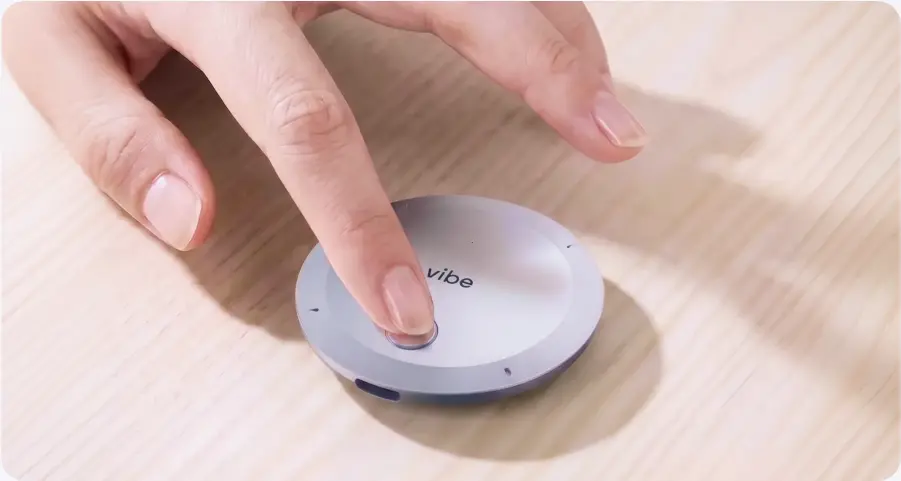 Finger pressing the button on a round Vibe Dot AI recorder placed on a wooden desk surface
